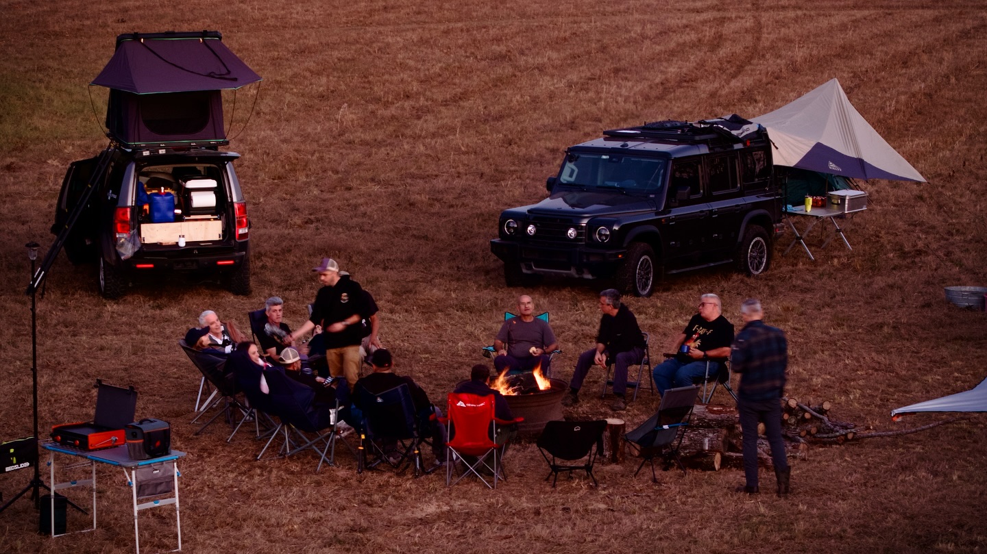 Overlanders gathered in camp chairs around a campfire at dusk with rooftop tents behind them